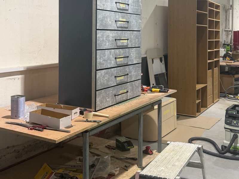 Grey chest of drawers on the work table, a shelving unit behind